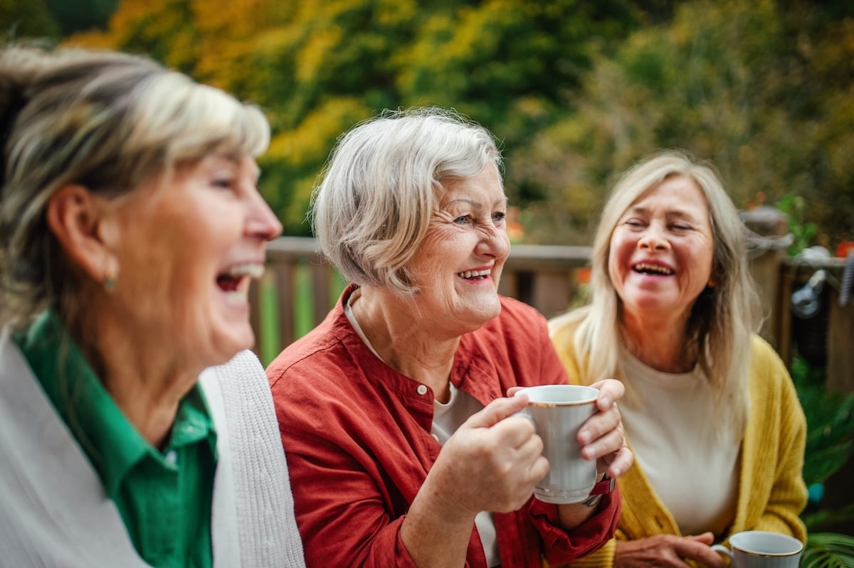 Three older women laughing with coffee outdoors