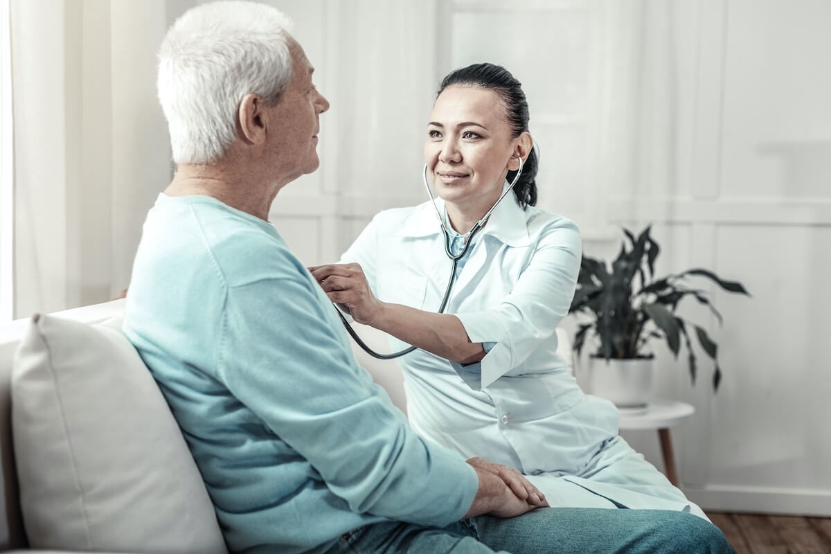 Nurse examining elderly man with stethoscope at home