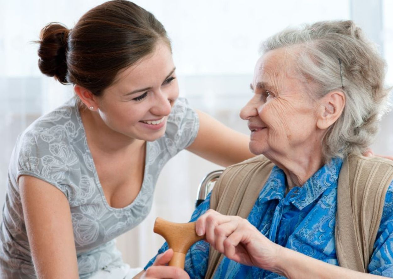 Young caregiver smiling at elderly woman with cane