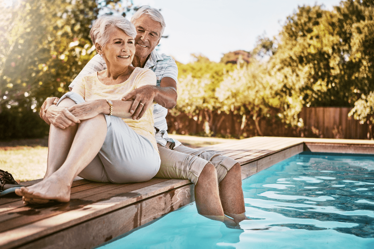 Happy elderly man and woman relaxing and hugging with feet in water by a poolside.