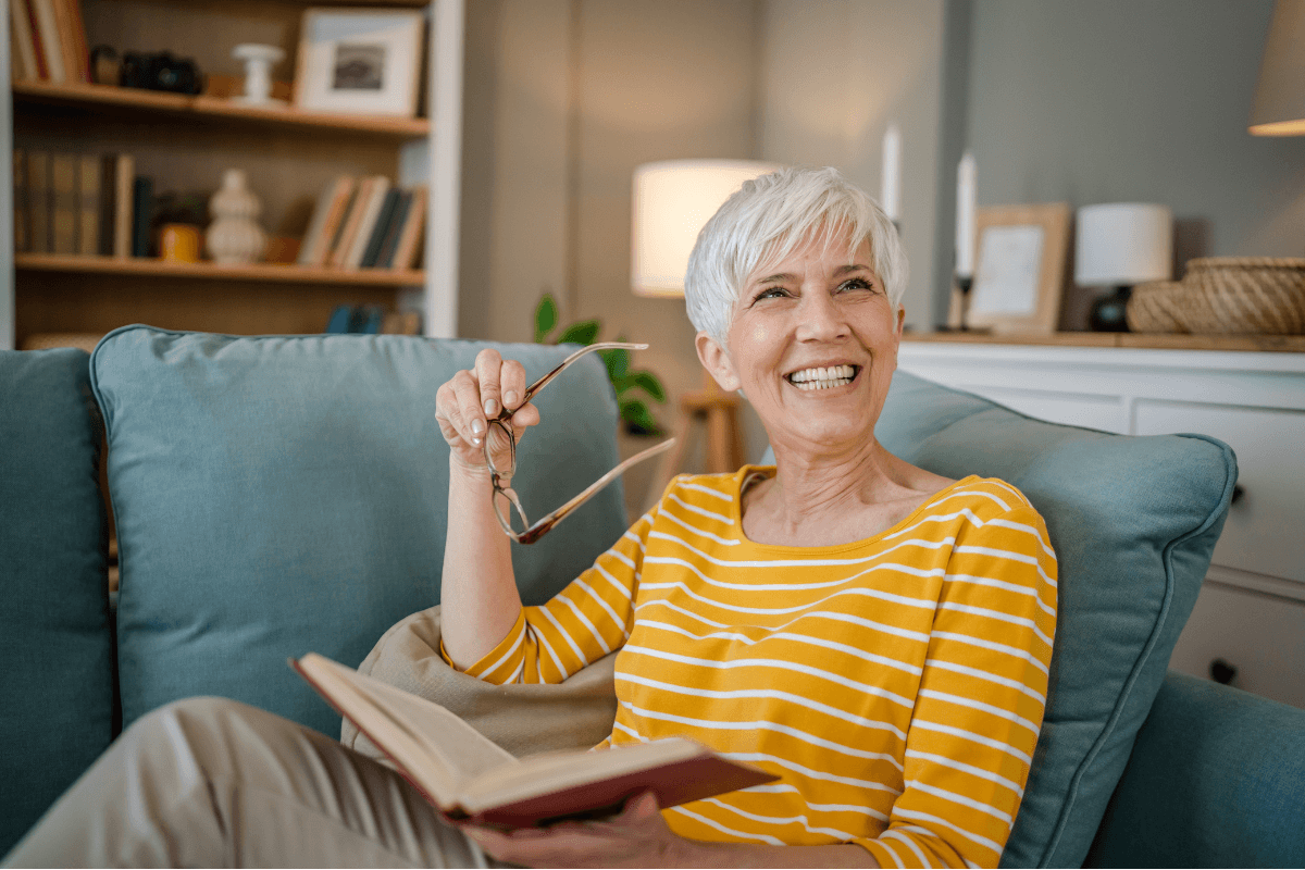 A mature, modern grandmother relaxes at home, reading a book with her glasses on.