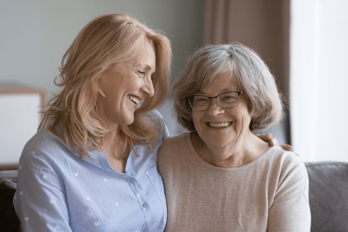 A cheerful elderly mother and her middle-aged daughter share a warm embrace on the couch at home.