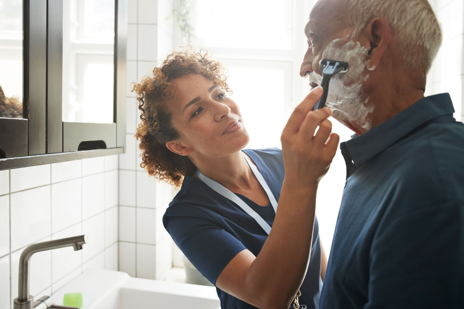 Healthcare worker shaving older man in a bathroom