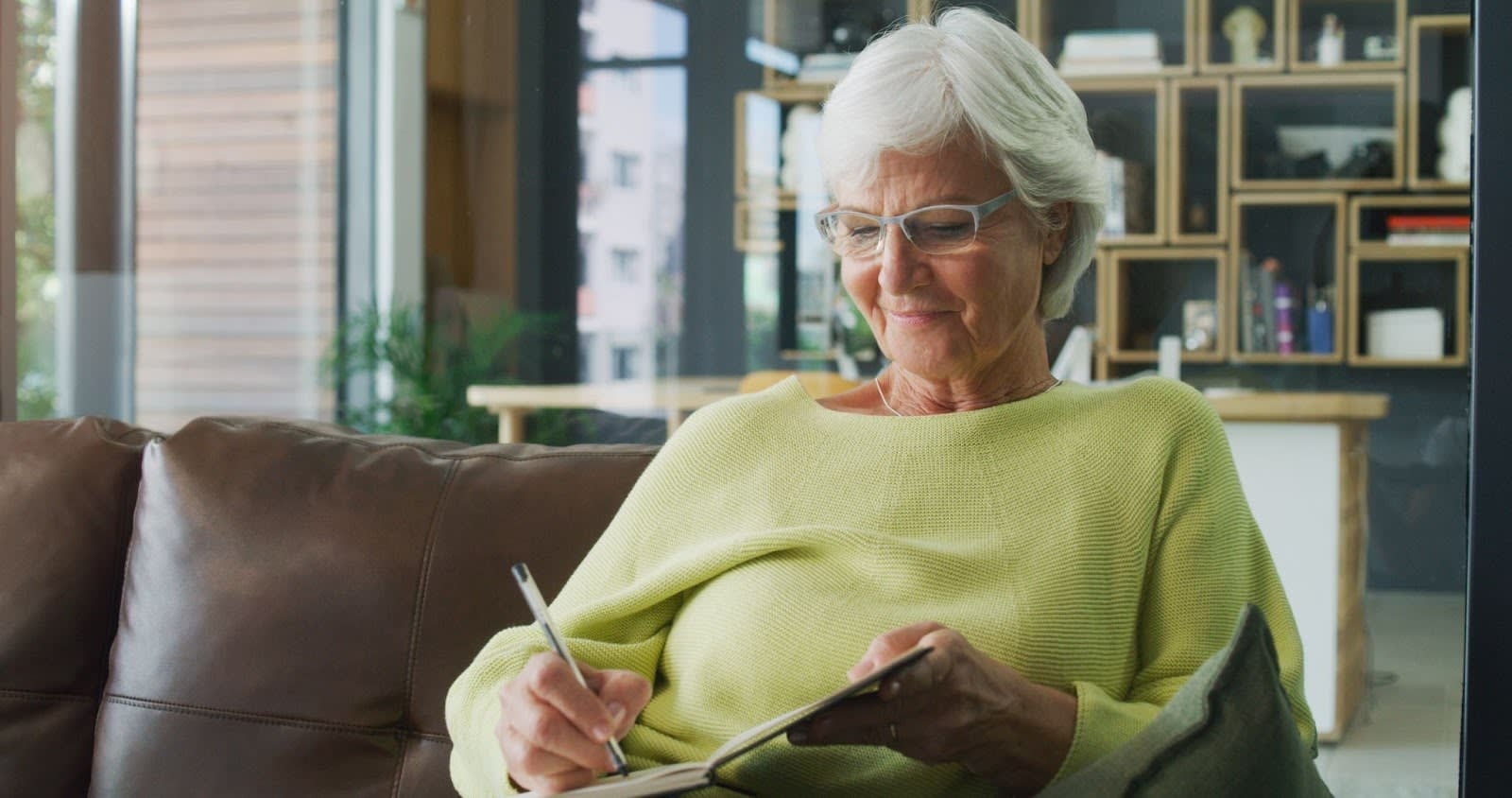 Older woman smiling and writing in a notebook while sitting on a couch