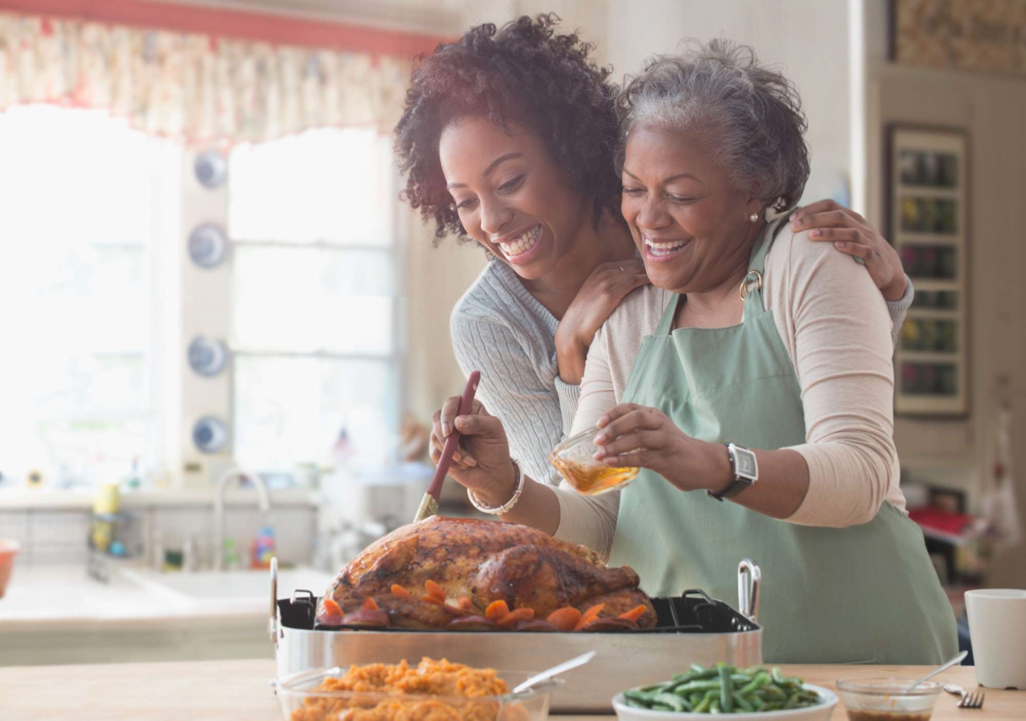 Younger woman and older woman smiling in a kitchen while basting a turkey