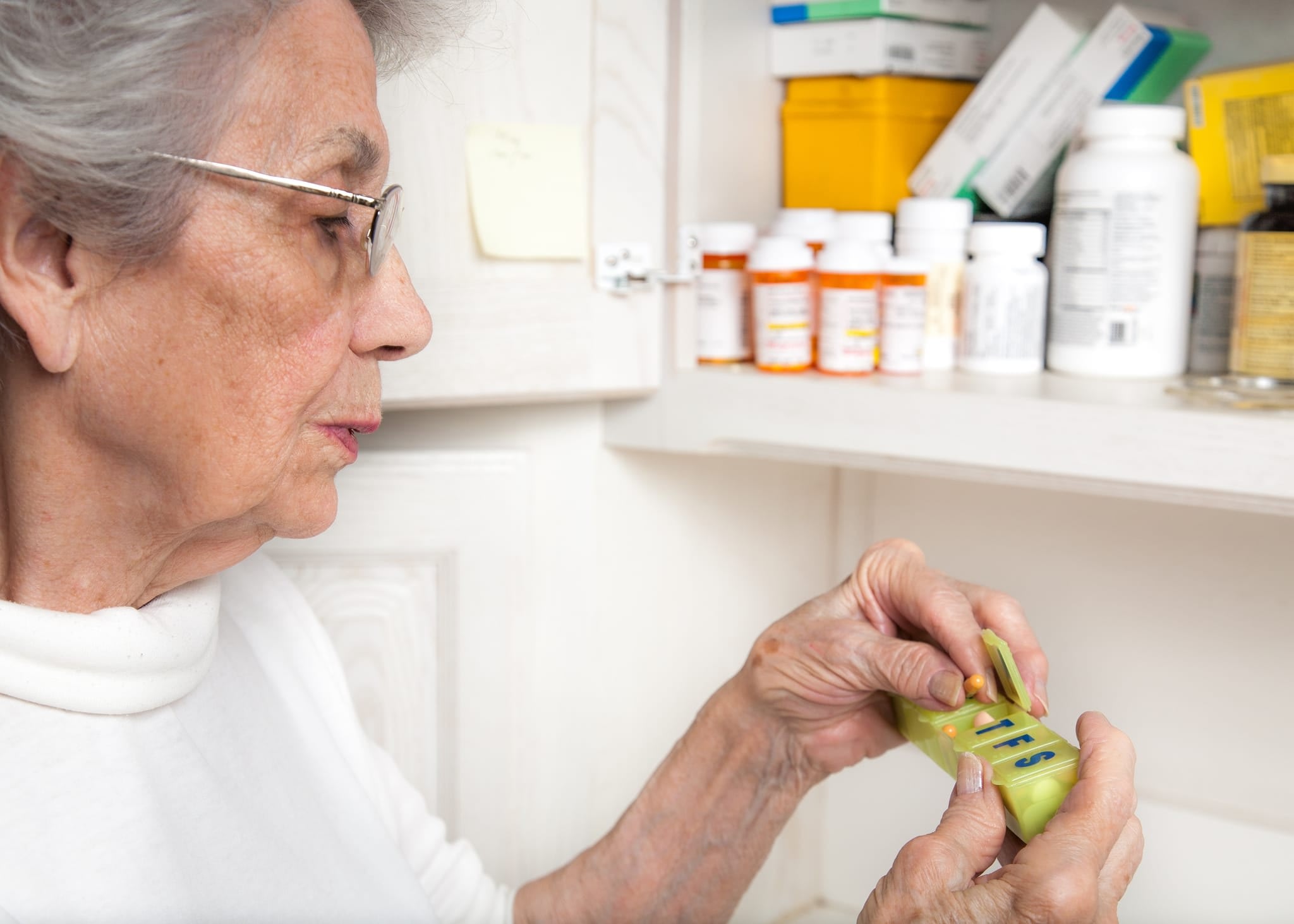 Senior woman standing in front of an open medicine cabinet placing pills into a daily pill container.
