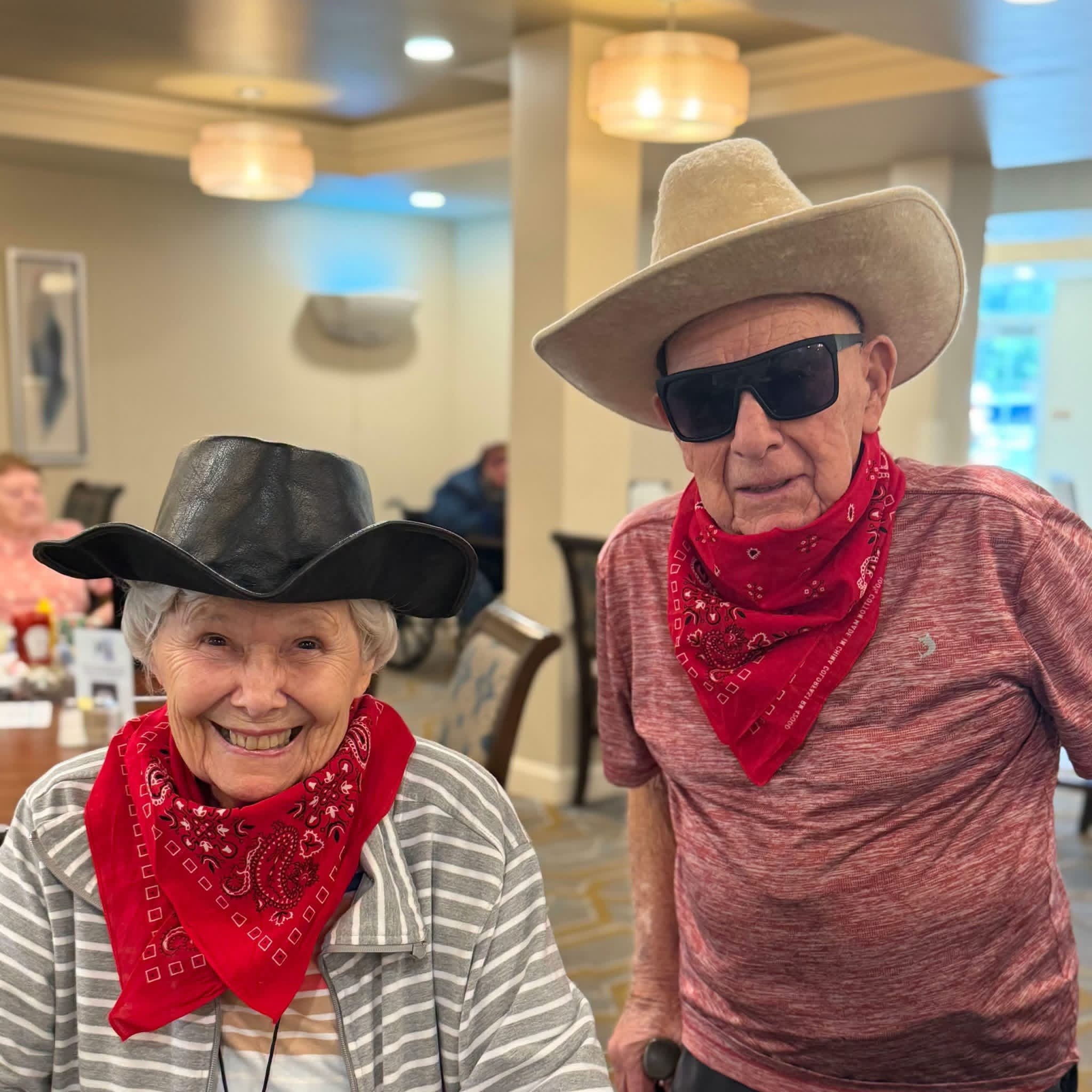 Two seniors wear cowboy hats and red bandanas indoors.