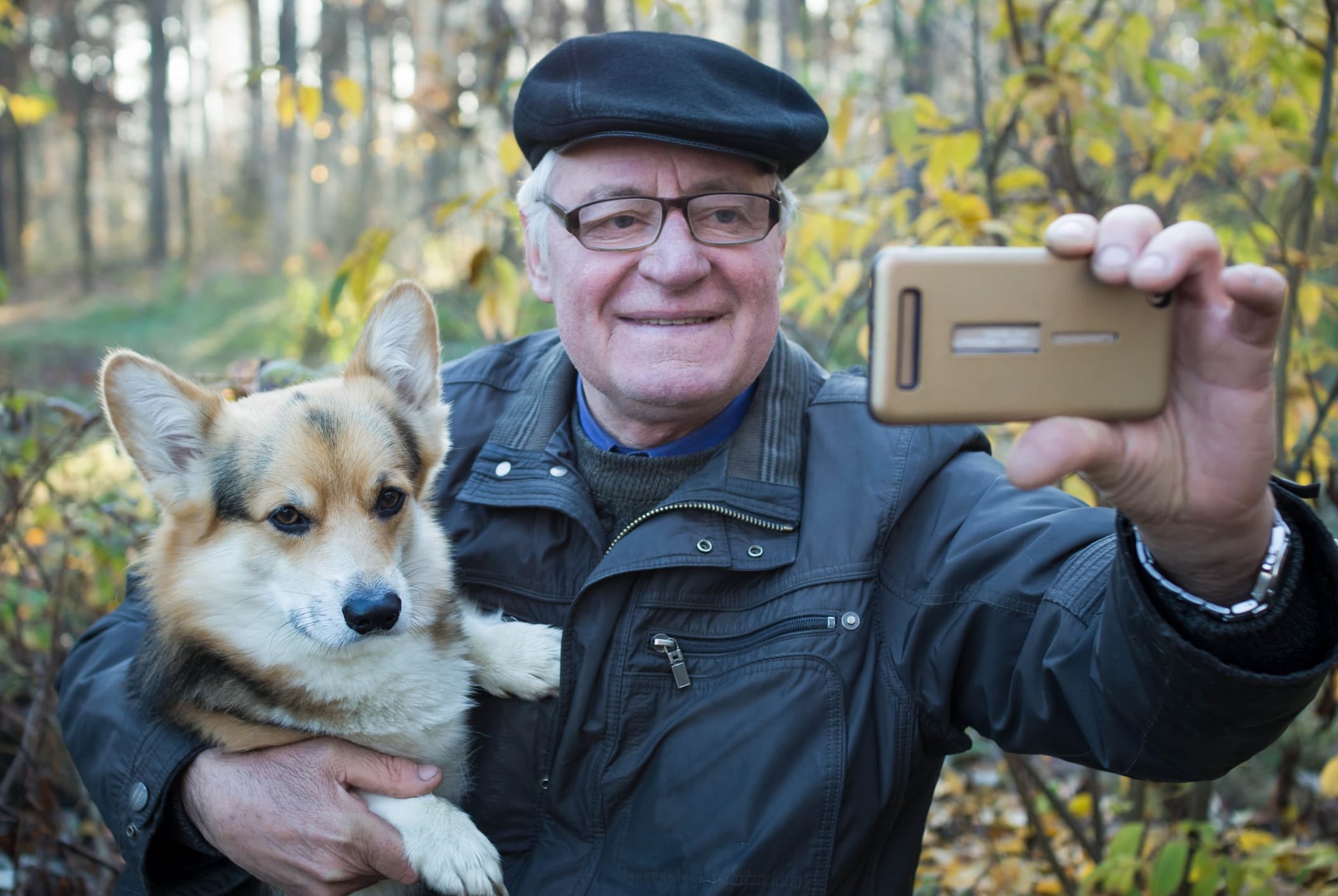An older man holding a Corgi dog takes a selfie with his phone in the forest.