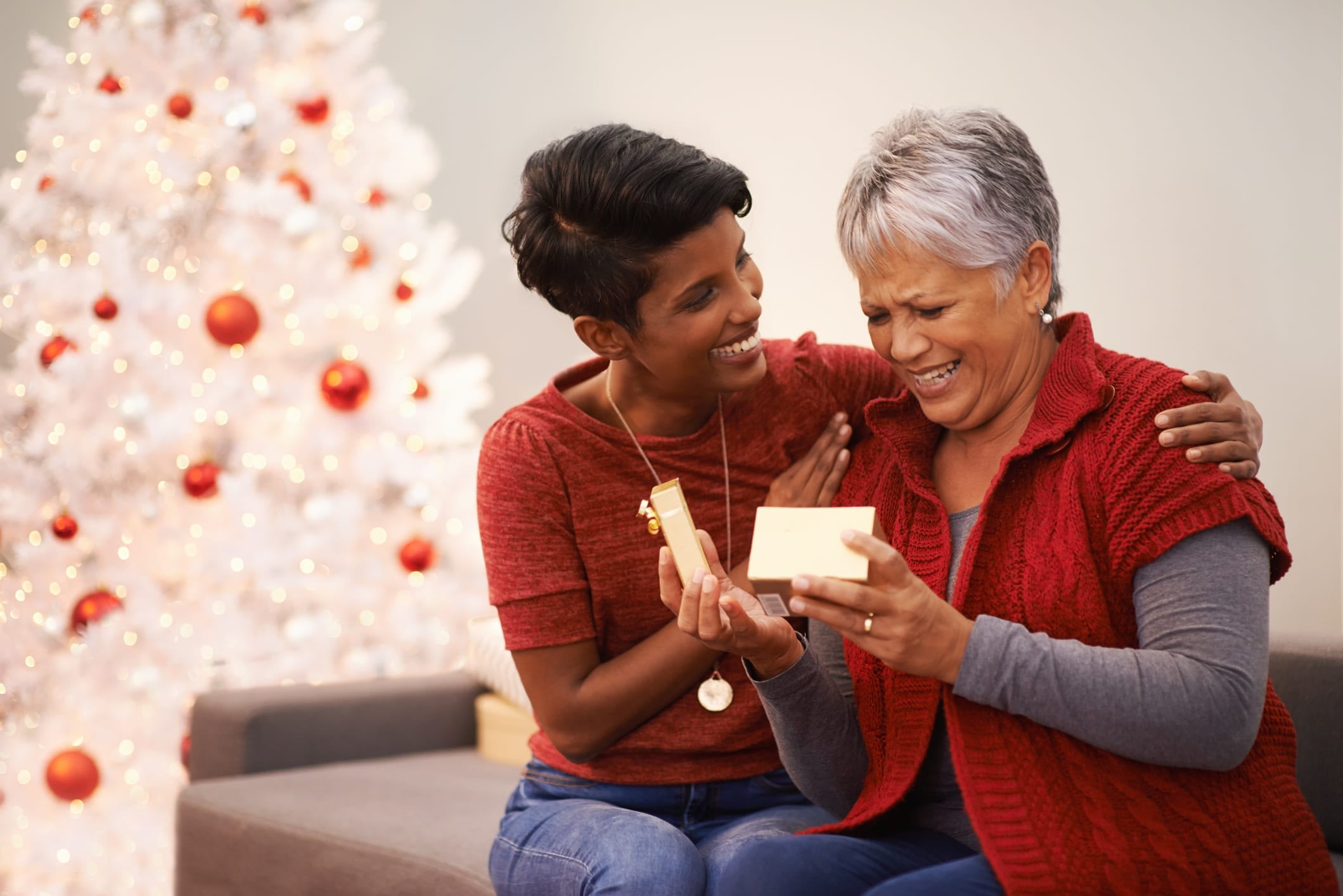 An adult daughter and her senior mother sitting on a couch. The daughter has her hands on her mother's shoulders as her mother admires a gift she just opened.