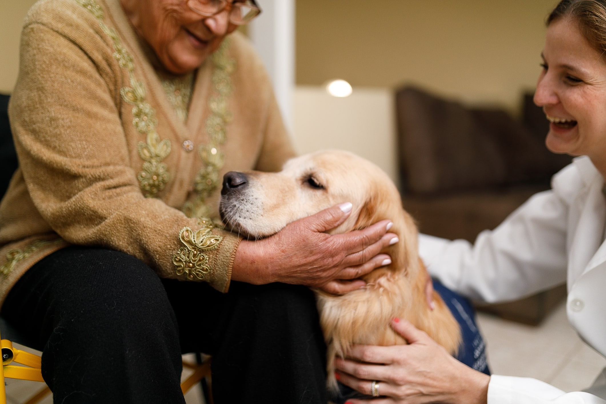 A smiling senior woman and her caregiver pet a therapy dog.
