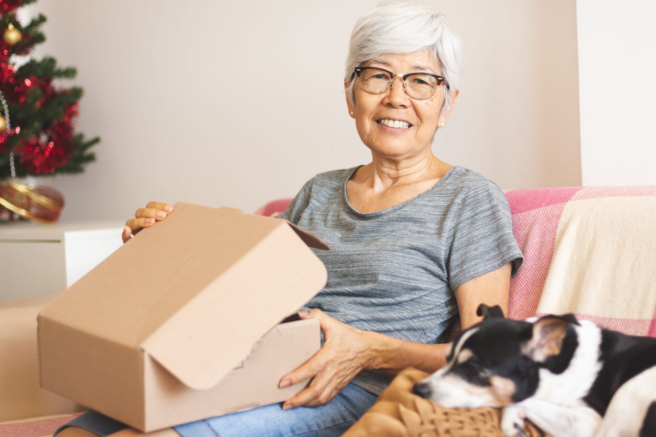Senior woman sitting on a couch next to a sleeping dog, opening a box, with a Christmas tree in the background.