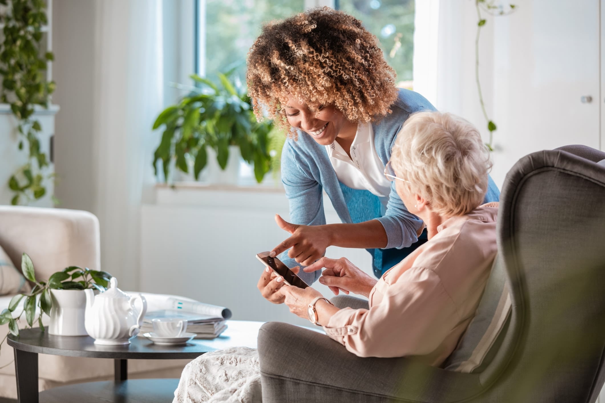 A young woman smiling and pointing at a smartphone while standing next to a seated older woman