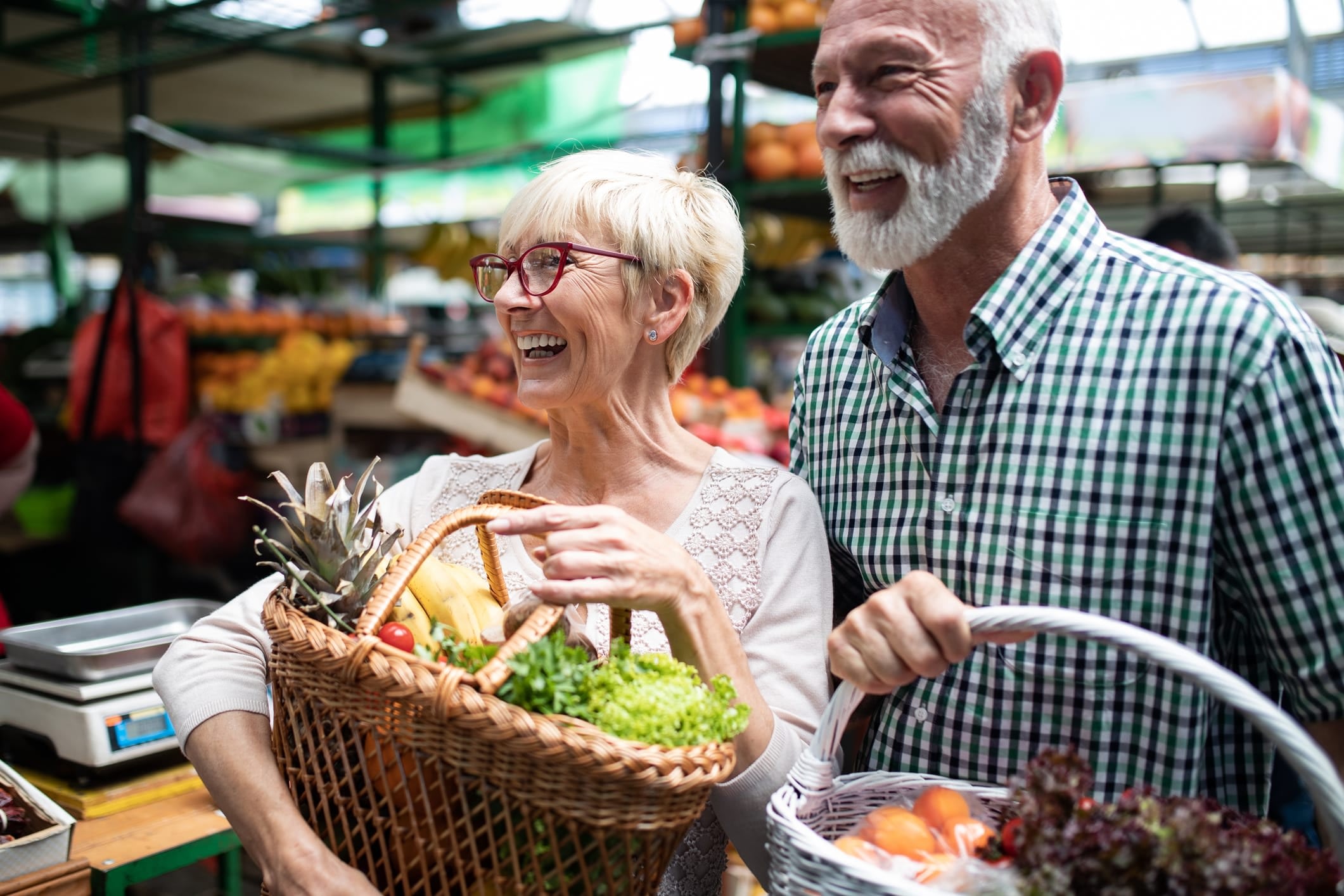 A smiling, older man and woman carry baskets of fruits and vegetables at a farmers market.