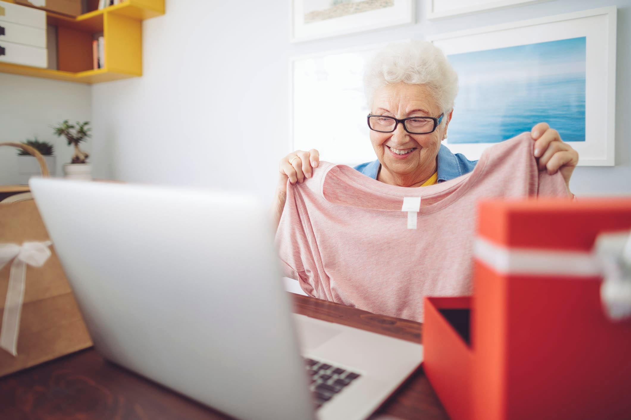 Senior woman holding up a shirt and smiling with an open box and open laptop computer in the foreground.