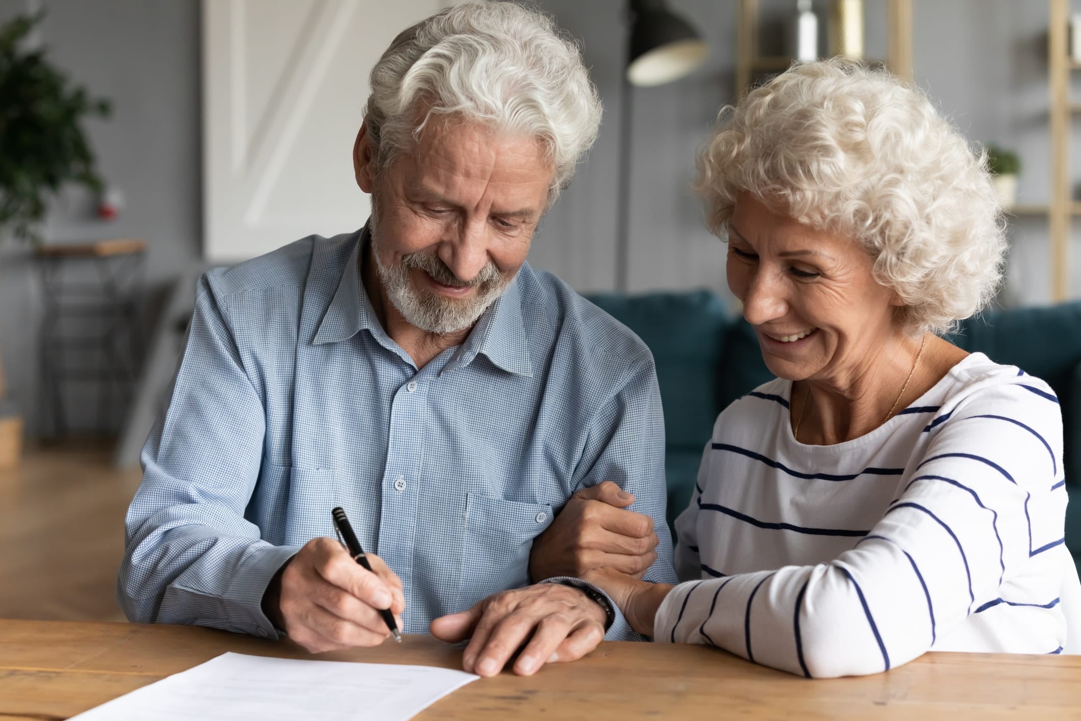 Senior man and woman sitting at a table, smiling, as man prepares to sign a paper.