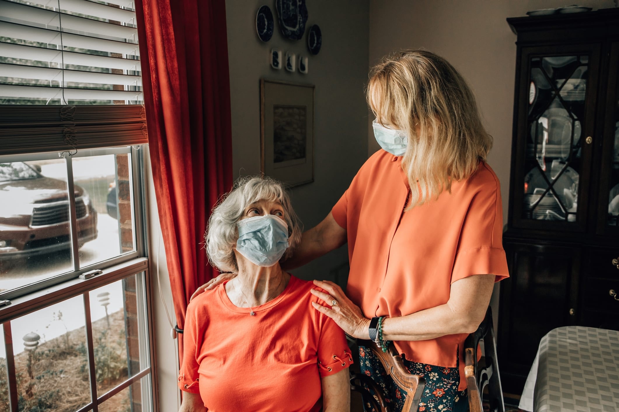 Senior woman with mask sitting in a chair looking up at younger woman who has her hands on the senior woman’s shoulders