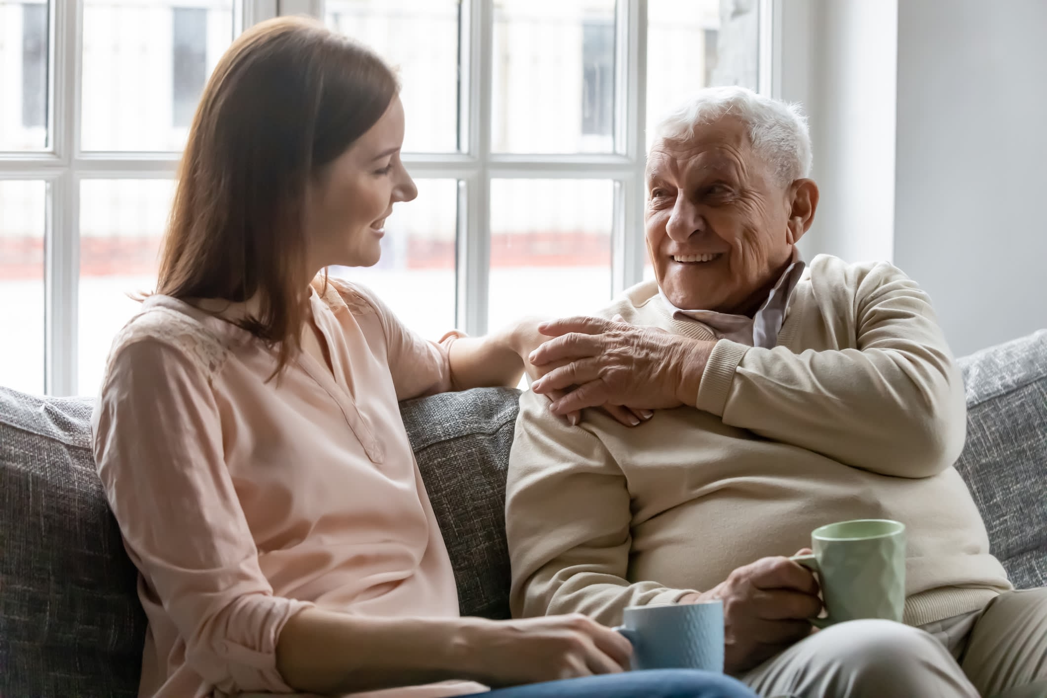 Young woman and senior man sitting on a couch, holding hands, and smiling at one another.