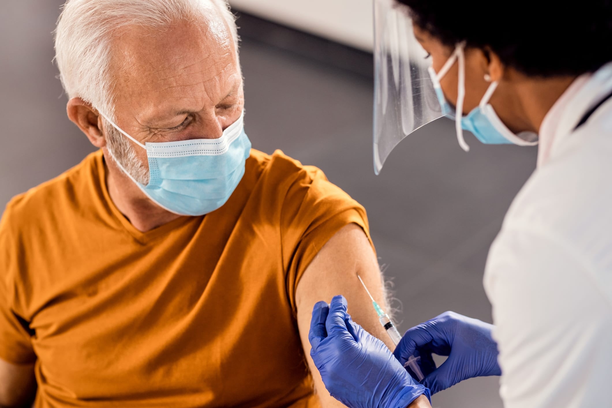 An older man wearing a face mask receiving a vaccine injection in his upper arm.