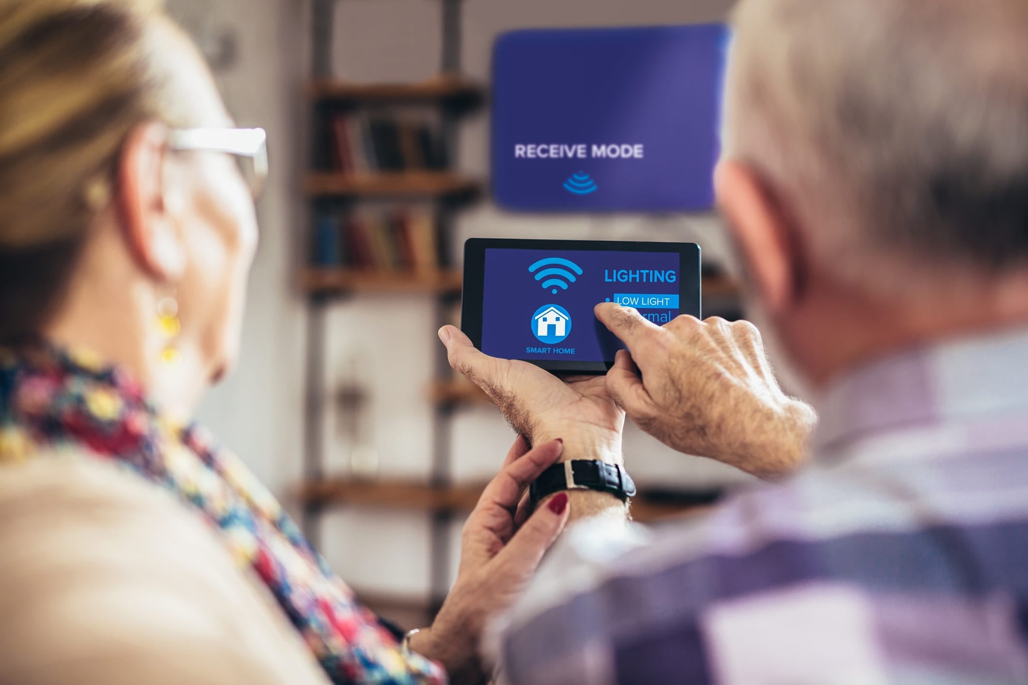 Senior man and woman holding a smartphone or small tablet computer and touching screen that shows an image of a house and the word “Lighting.”