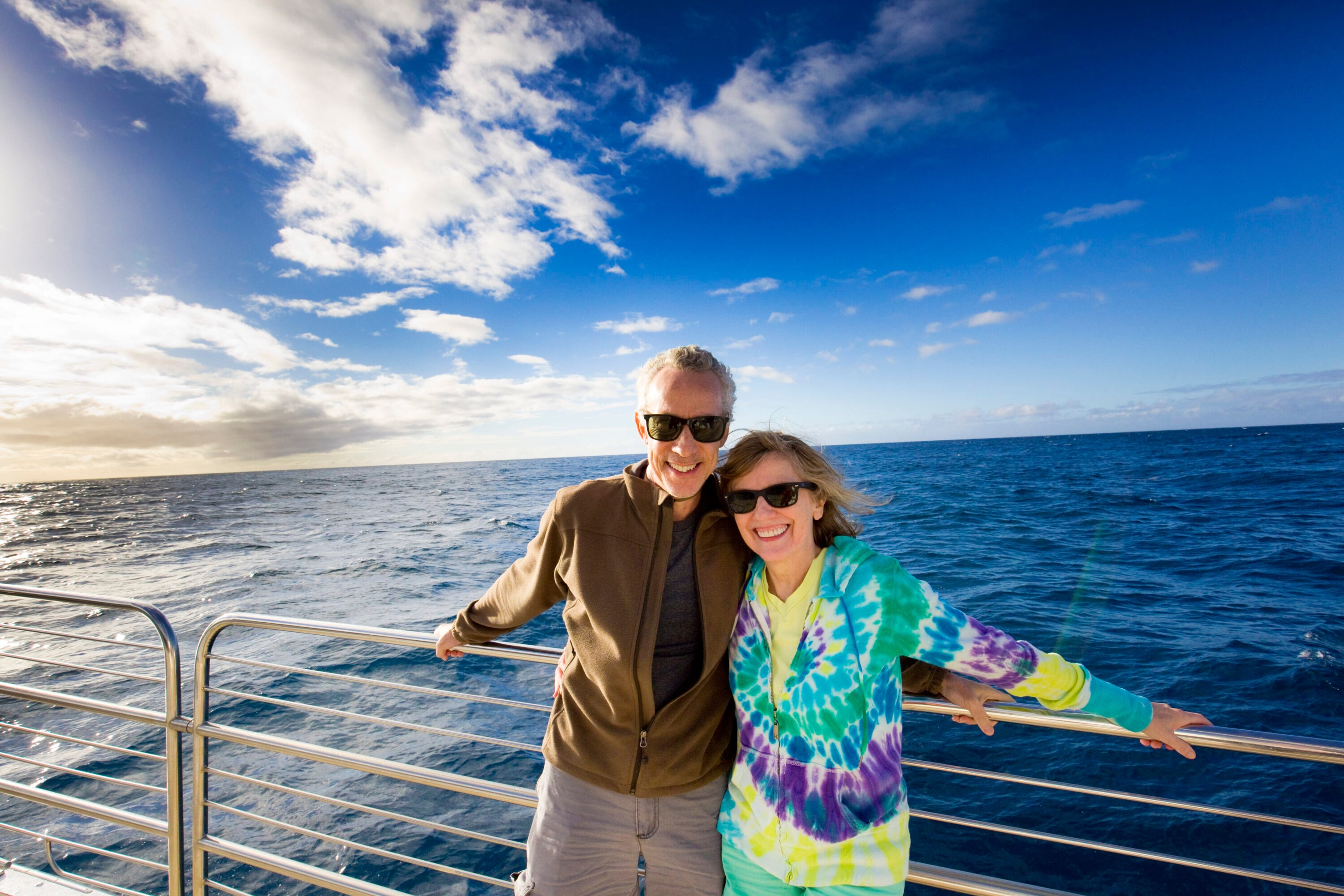 A senior man and woman leaning against the rails on a ship smiling with the ocean in the background.